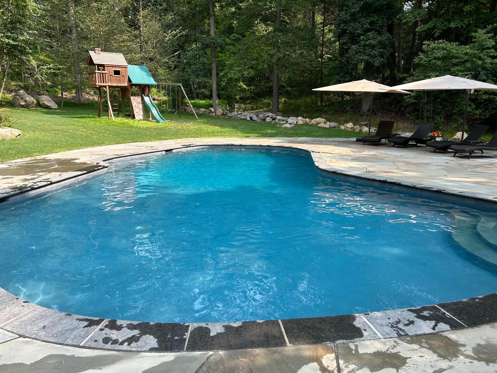 Free-form pool with flagstone patio, umbrellas, and a children's playset in the background
