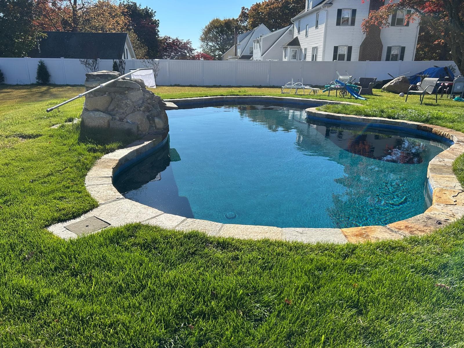 Kidney pool with a natural rock waterfall feature, white vinyl fence, and fall foliage