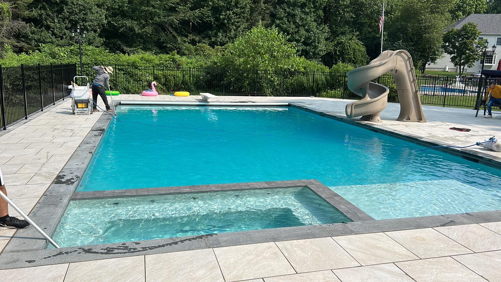 Rectangular pool with attached spa, slide, and paver deck being serviced by a crew member